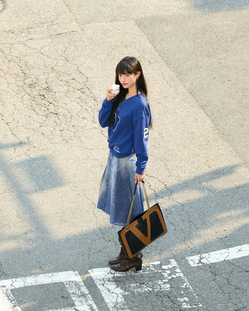 Woman in a blue sweatshirt and denim skirt standing in sunlight on a city street